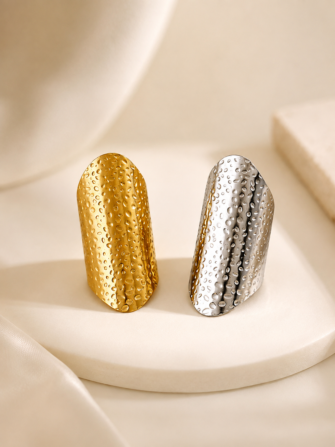 Two sculptural, hammered metal rings, one in polished gold and one in polished silver, displayed side-by-side on a round cream-colored stone pedestal.