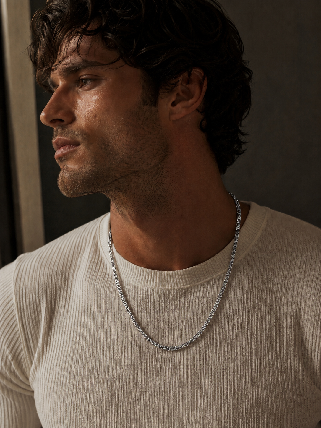 A close-up of a man wearing a silver chain necklace over a cream ribbed t-shirt, set against a dark, moody background.