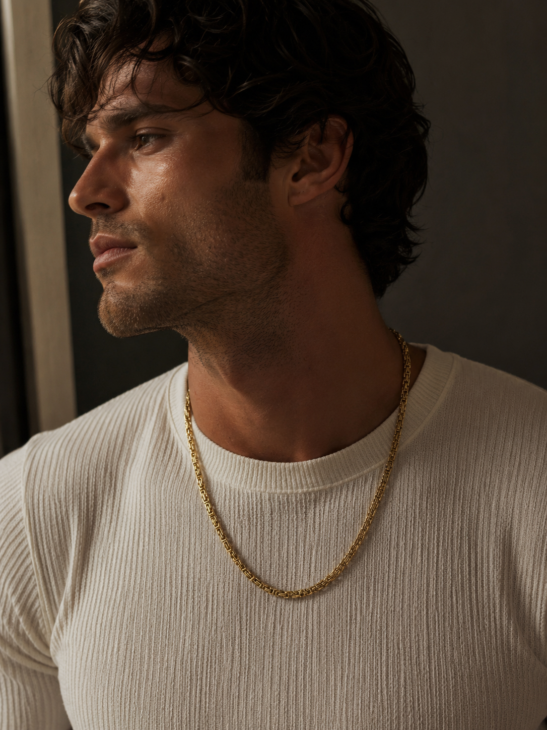 A man in profile wearing a textured off-white ribbed crewneck shirt and a gold chain necklace, set against a dark, moody background.