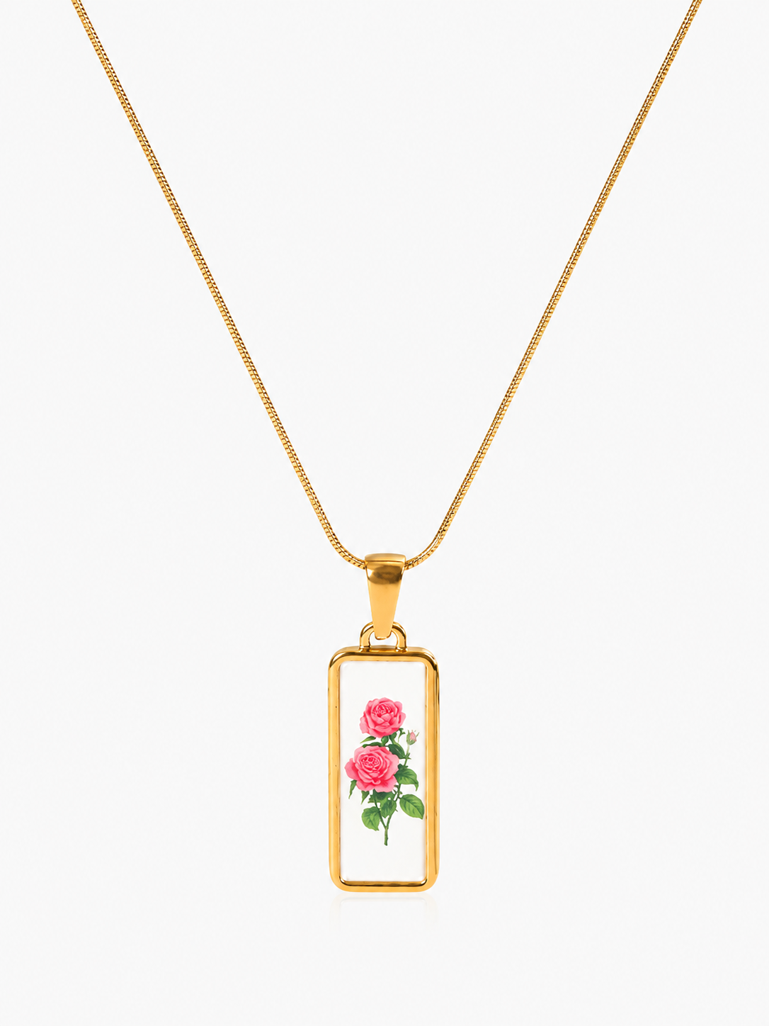 A gold rectangular pendant necklace featuring an illustration of pink roses with green leaves on a white background, suspended from a gold chain against a light gray studio backdrop.