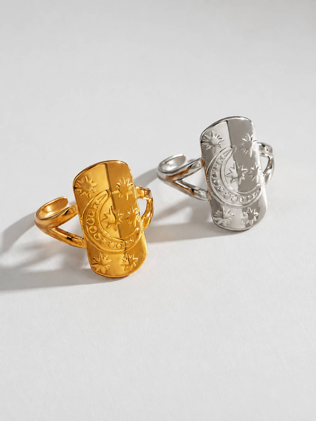 Two engraved cuff-style rings on a light gray background, one in polished gold and one in polished silver, each with a shield-like face and decorative border details.