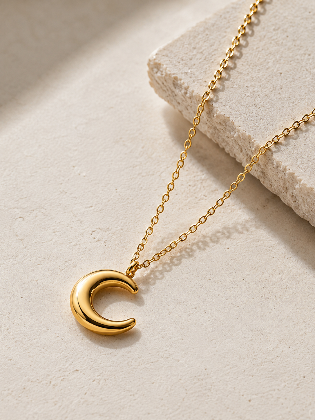 A polished gold crescent moon pendant necklace on a delicate gold chain, styled on a textured beige stone slab against a neutral beige background.