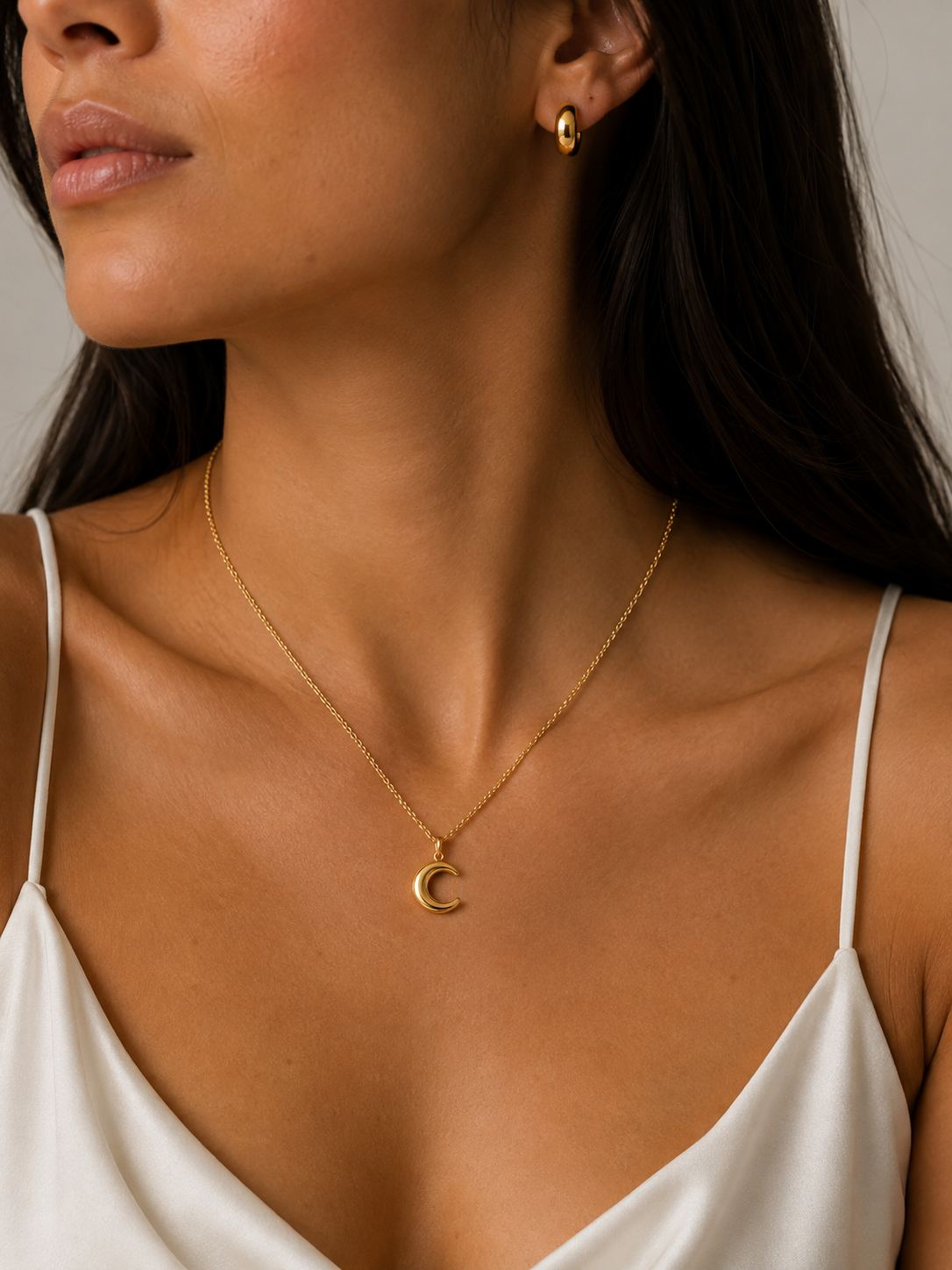 A close-up of a woman wearing a delicate gold chain necklace with a crescent moon pendant, paired with a small gold hoop earring and a white camisole top.