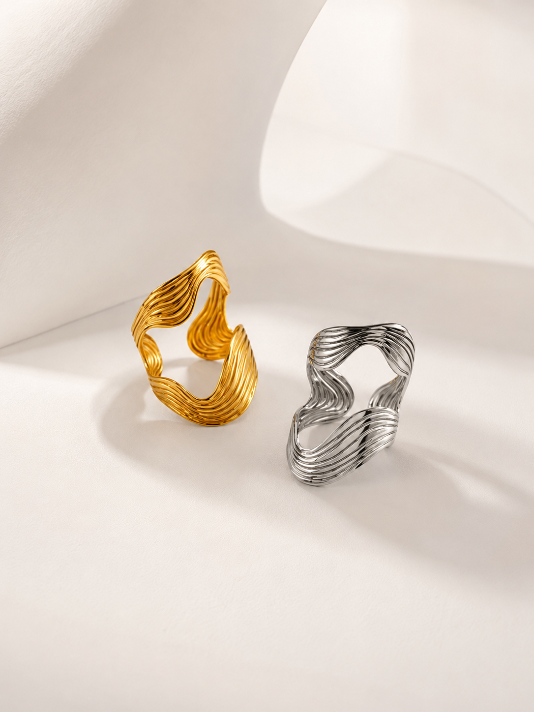 Two twisted open-band rings with a wavy, layered design, one in polished yellow gold and the other in silver, displayed on a clean white surface with soft shadows.